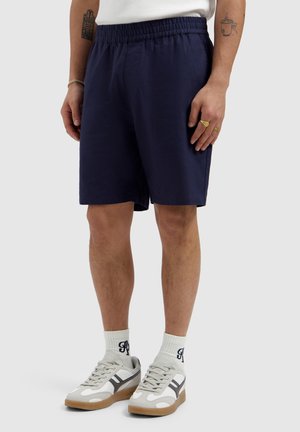 Mann trägt marineblaue Shorts mit elastischem Bund, weiße Socken mit schwarzer Schrift, weiße und graue Sneaker sowie Ringe an der rechten Hand mit Tätowierungen an den Armen.