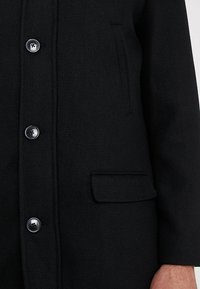 Černý kabát s texturovanou látkou, pěti velkými knoflíky a postranní kapsou na levé straně, s upraveným střihem.