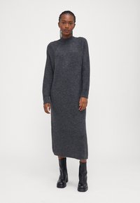 Μακριά γκρι πλεκτή φορεσιά με ψηλό γιακά και ριχτές μανσέτες, διαθέτει ευθεία σιλουέτα και πλαϊνές σχισμές, συνδυασμένη με μαύρες μπότες έως τον αστράγαλο.