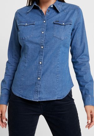 Camicia di denim blu con maniche lunghe, colletto a punta, due tasche sul petto e bottoni a pressione. Presenta un design aderente e orlo curvo.