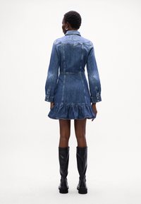 Vestido de ganga com mangas longas, bainha com pregas e fecho de botão, apresentando uma cintura ajustada e lavagem azul clara. Com botas pretas até ao joelho.