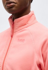 Κοντινό πλάνο ατόμου που φοράει ανοιχτό ροζ φλις μπουφάν με φερμουάρ, με το ανάγλυφο λογότυπο HH στο στήθος, σε απλό ανοιχτό φόντο.