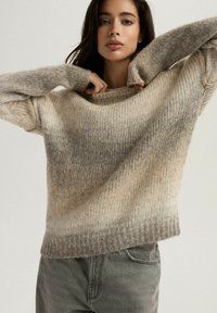 Sweter dzianinowy w gradacji beżowych i szarych odcieni, z luźnym dekoltem, ściągaczami na rękawach i miękką teksturą. W połączeniu z szarymi spodniami.