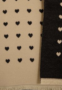 Écharpe en mélange de laine avec un côté beige orné de motifs de cœurs noirs répétés et un côté noir contrastant. Finitions avec des bords à franges.