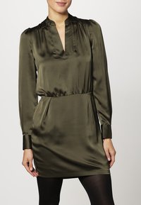 Abito in satin verde oliva con maniche lunghe, scollo a V e vita elastica. Presenta una texture liscia e un dettaglio di spalla leggermente arricciato.