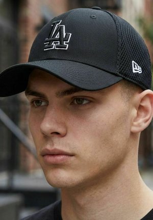 Junger Mann mit blasser Haut, der eine schwarze LA-Baseballkappe mit Netzrücken trägt und mit neutralem Gesichtsausdruck nach vorne schaut, in einer städtischen Umgebung im Freien.