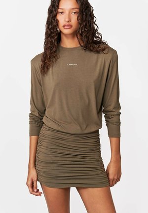 Kobieta o kręconych włosach, ubrana w oliwkowo-zieloną mini sukienkę na długi rękaw z marszczeniami i logo "LAMANIA" na piersi, stojąca na białym tle.