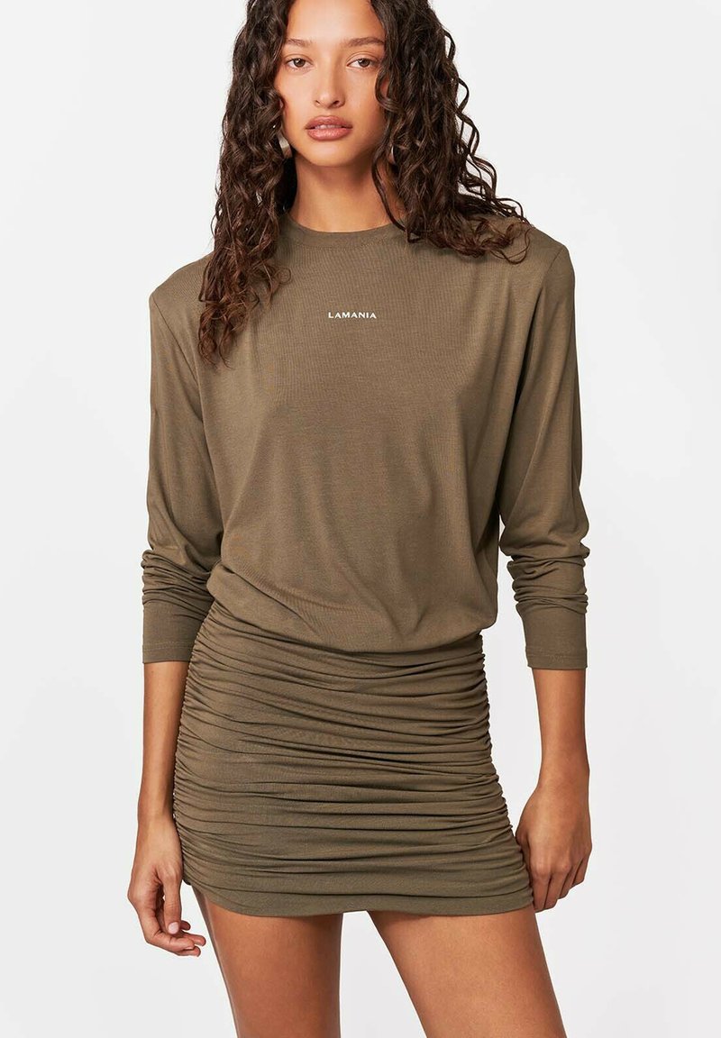 Kobieta o kręconych włosach, ubrana w oliwkowo-zieloną mini sukienkę na długi rękaw z marszczeniami i logo "LAMANIA" na piersi, stojąca na białym tle.