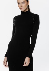 Czarna dzianinowa sukienka z wysokim kołnierzem, prążkowaną strukturą, długimi rękawami oraz dekoracyjnymi czarnymi guzikami na lewym ramieniu. Dopasowany krój.