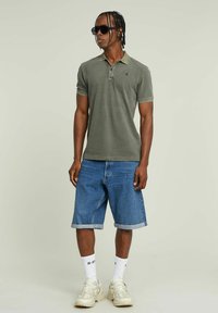 Oliwkowa koszulka polo z krótkim rękawem, kontrastowym kołnierzykiem, w zestawie z dżinsowymi szortami i jasnymi sneakersami. Model nosi okulary przeciwsłoneczne.