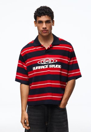 Ung mand med krøllet hår iført en rød og marineblåstribet poloshirt med teksten "SURFACE STUDIO", stående med den ene hånd i lommen.