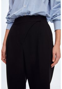 Personne portant un pantalon noir taille haute ajusté et une chemise à rayures bleues et blanches à boutons, les manches retroussées, se tenant avec les bras le long du corps.