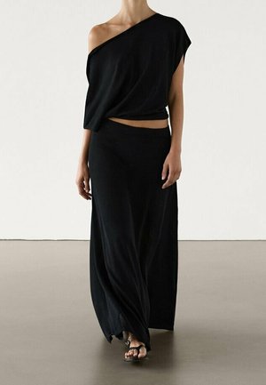 Woman wearing a black one-shoulder top and long skirt walking barefoot on a smooth floor against a plain white wall.