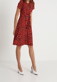 Czerwona sukienka w czarno-białe wzory lamparta, z krótkimi rękawami i wiązaniem w pasie. Połączona z białymi szpilkami.