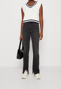Chaleco blanco de punto con detalles en azul marino, combinado con jeans negros acampanados con aberturas laterales, complementado con zapatillas blancas y un bolso negro.