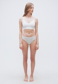 Νεαρή γυναίκα όρθια, ξυπόλητη, με λευκό αθλητικό σουτιέν και ανοιχτόχρωμο γκρι εσώρουχο, κοιτάζει προς τα αριστερά της μπροστά σε απλό φόντο.