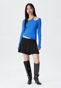 Eine Frau in einem leuchtend blauen asymmetrischen Langarmtop, einem schwarzen Minirock mit gefalteten Paneelen und kniehohen schwarzen Lederstiefeln, steht vor einem weißen Hintergrund.