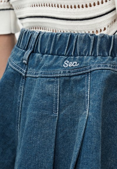 Κοντινό της μέσης μιας μπλε τζιν φούστας με ελαστική λεπτομέρεια και λευκή κεντημένη λέξη "Sea", φορεμένη με λευκό πλεκτό τοπ.