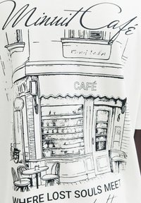 Bijela majica s crnim crtežom kafića sa prugastim tendom, vanjskim sjedenjem i natpisom "Minuit Café" te "GDJE SE SUSRETAJU IZGUBLJENE DUŠE."