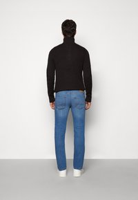 Man som står vänd mot en vägg och bär en svart polotröja, blå jeans och vita sneakers på ett enfärgat grått golv.