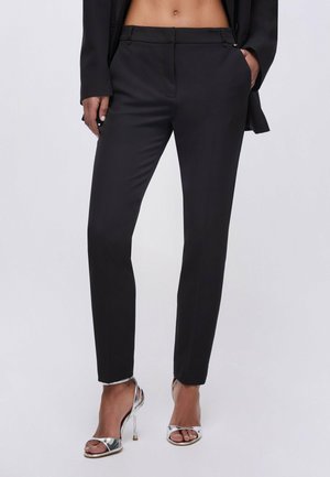 Pantaloni neri su misura a taglio slim indossati con sandali argentati con tacco alto, mostrati dalla vita ai piedi su uno sfondo semplice.