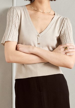 Femme les bras croisés portant un haut en maille beige à col en V avec des manches courtes à motifs, un collier doré, une bague en or et un pantalon noir.
