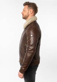 Giacca in pelle marrone con un colletto beige in peluche, polsini a coste e fascia in vita. Presenta tasche laterali e un piccolo emblema sulla manica.