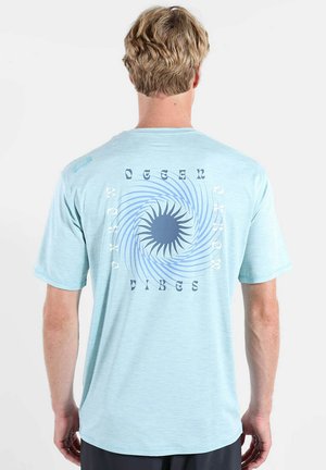 Homme aux cheveux blonds porte un t-shirt bleu clair avec un motif de soleil et spirale bleu foncé, texte "Ocean Vibes" dans le dos sur fond blanc.