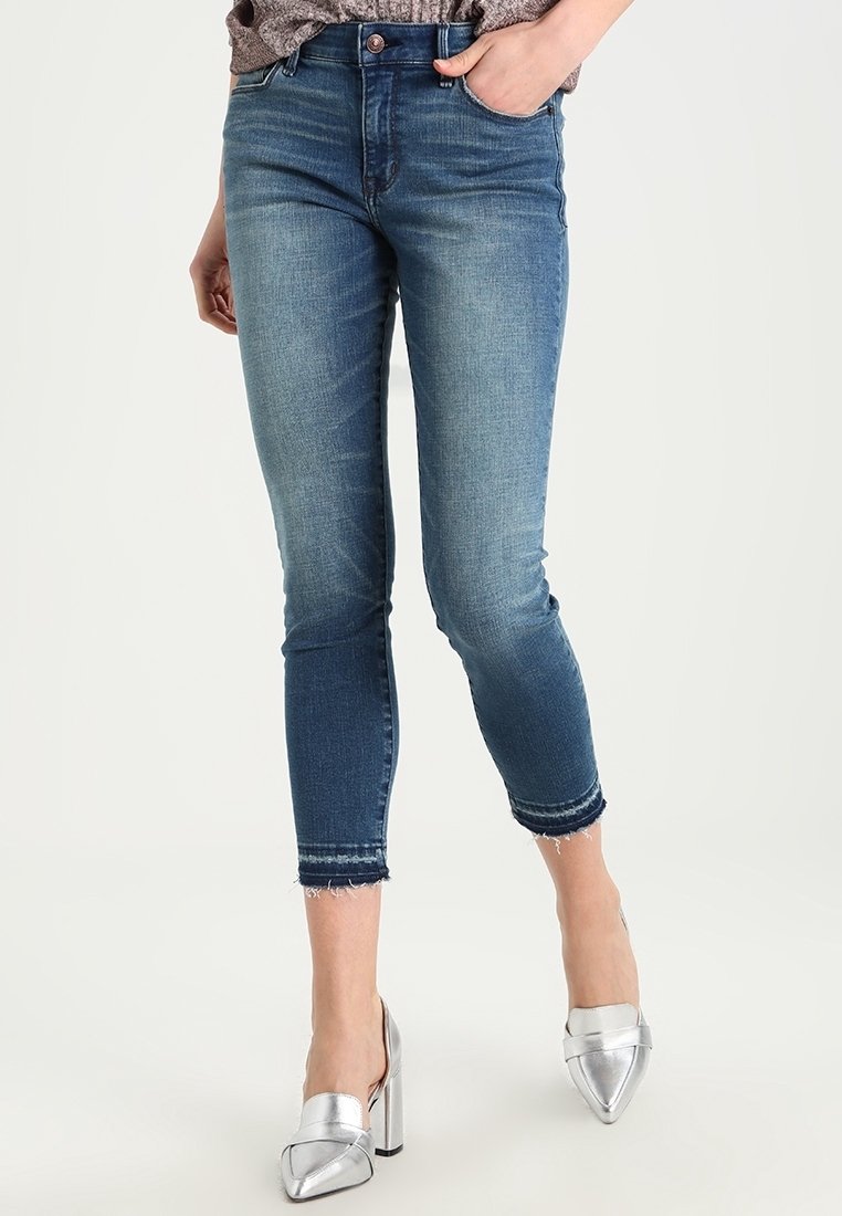 Pantalones vaqueros de mezclilla azul desgastados con un diseño ajustado, longitud recortada y dobladillo deshilachado; combinados con zapatos de tacón alto plateados.