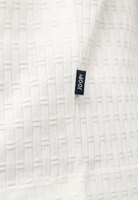 Tissu blanc texturé avec un motif côtelé vertical et horizontal et une petite étiquette noire portant la mention "JOOP!" attachée à la couture.
