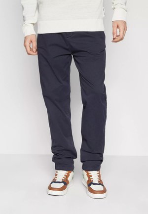 Chinos blu navy in cotone, con vestibilità dritta, dotati di tasche laterali e passanti per cintura, abbinati a sneaker bianche e marroni.