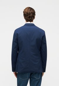 Άνδρας με  καστανά σγουρά μαλλιά, ντυμένος με μπλε σκούρο σακάκι και μπλε τζιν, στέκεται αντικρίζοντας ένα απλό λευκό φόντο.