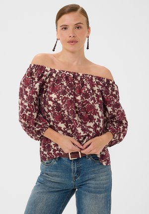 Femme portant un haut épaules dénudées avec un motif floral rouge et un jean bleu, debout les mains près de la taille sur un fond uni.