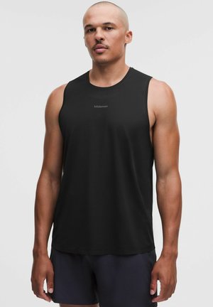 Atletisk mand iført en sort ærmeløs Lululemon-trøje og mørke shorts står mod en ensfarvet lys baggrund.