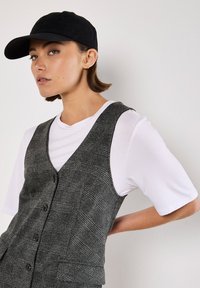 Gorra de béisbol negra, camiseta blanca y chaleco de cuadros gris con botones y bolsillos. Primer plano de un modelo contra un fondo neutro.