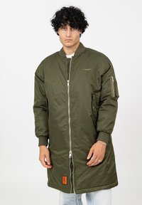 Man met krullend zwart haar in een lange olijfgroene bomberjack met een rits aan de voorkant en een patch, staande tegen een plain witte achtergrond.