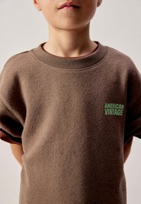 Bambino che indossa una felpa marrone a maniche corte con la scritta "American Vintage" in verde sul petto, in piedi davanti a uno sfondo neutro.