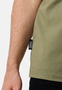 Oliven grøn bomuld T-shirt med en rund kant og et sort mærke syet på siden nær en bar arm. Teksturen ser glat og blød ud.