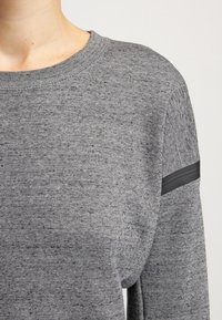 Grå tröja i strukturerad stickning, med rund halsringning och ett horisontellt svart rand på ärmen som accent.