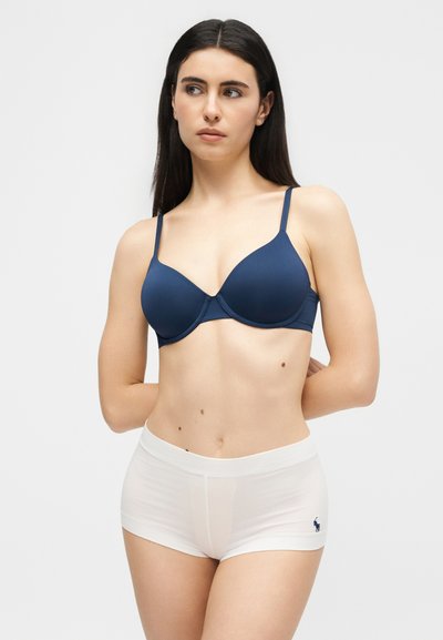 Γυναίκα με μακριά σκούρα μαλλιά, φοράει μπλε σουτιέν και λευκά σορτς εσώρουχα, στέκεται με τα χέρια πίσω από την πλάτη, κοιτάζοντας προς τα πλάγια.