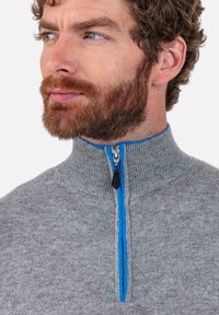 Pull gris à fermeture éclair avec un col haut, une fermeture éclair bleue contrastante, une texture côtelée et une coupe ajustée. Fabriqué en tissu doux.