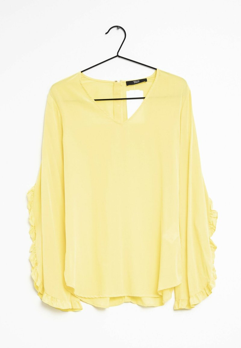 Blouse jaune avec des manches longues et volantes. Décolleté en V et fente en forme de trou de serrure au dos. Tissu léger qui tombe doucement sur le corps.