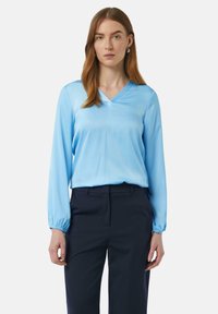 Himmelbluse aus Seide mit V-Ausschnitt und langen Ärmeln, die an den Handgelenken gerafft sind, kombiniert mit dunkelblauen, taillierten Hosen.