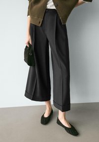 Schwarze Hosen mit weitem Bein und umgeschlagenem Saum, kombiniert mit dunkelgrünen, flachen, plissierten Schuhen. In einer Hand wird eine strukturierte, dunkle Handtasche gehalten.