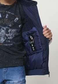 Dieťa oblečené v tmavom grafickom tričku a modrých džínsoch, ktoré drží otvorenú námorníckom modrú vypaľovanú bundu so soft inner lining a viditeľnou označení o starostlivosti.