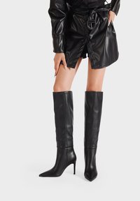 Stivali neri al ginocchio realizzati in pelle liscia, con punta affilata e tacco a spillo, dal profilo aderente.