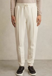 Pantaloni de trening de culoare crem, cu un elastic la talie și șnur, față pliată și manșete cu nervuri, asortați cu pantofi sport negri. Textură netedă.