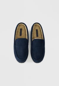Tmavě modré semišové mokasíny s měkkou béžovou umělou kožešinou uvnitř a ozdobným prošíváním podél okrajů.