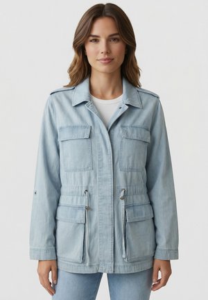 Femme portant une veste en denim bleu clair avec quatre poches à rabat à l'avant et une taille ajustable par cordon, se tenant devant un fond uni.