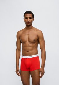 Κόκκινα βαμβακερά ανδρικά εσώρουχα-βοξέρ με λευκή μέση λογότυπου, με εφαρμοστή γραμμή και λεία υφή, παρουσιασμένα από ένα άτομο χωρίς μπλούζα.